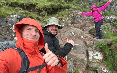 Three members of D&A staff climbing a steep path on a mountain and smiling