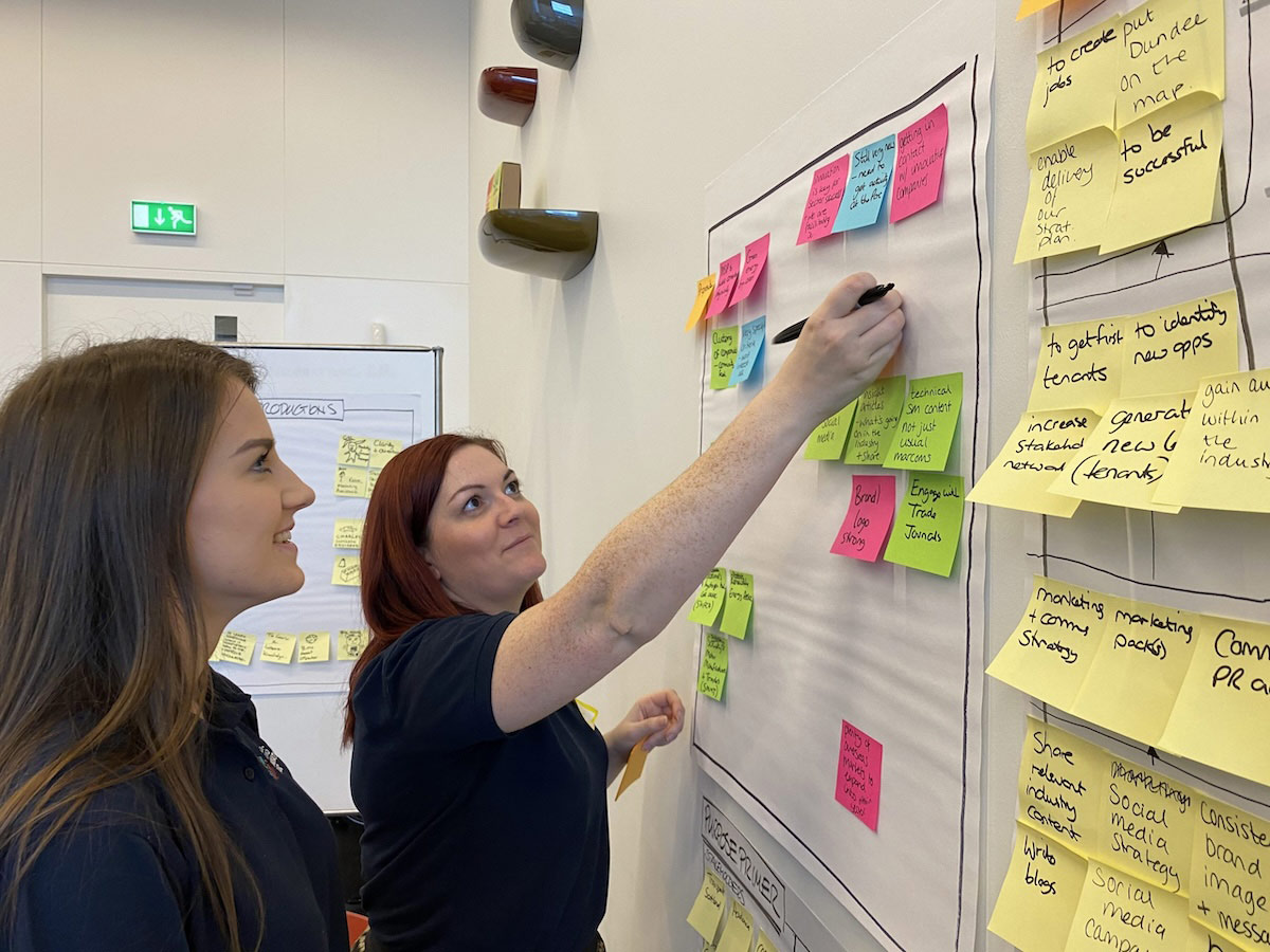 Two women from the Michelin Scotland Innovation Parc team are looking at a wall with post it notes stuck on it. One of the women has a pen in her hand and is sticking a post it note to the wall. 