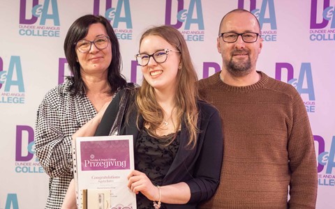 Student standing in the middle of parents holding certificate and prize in front of Dundee and Angus College backdrop