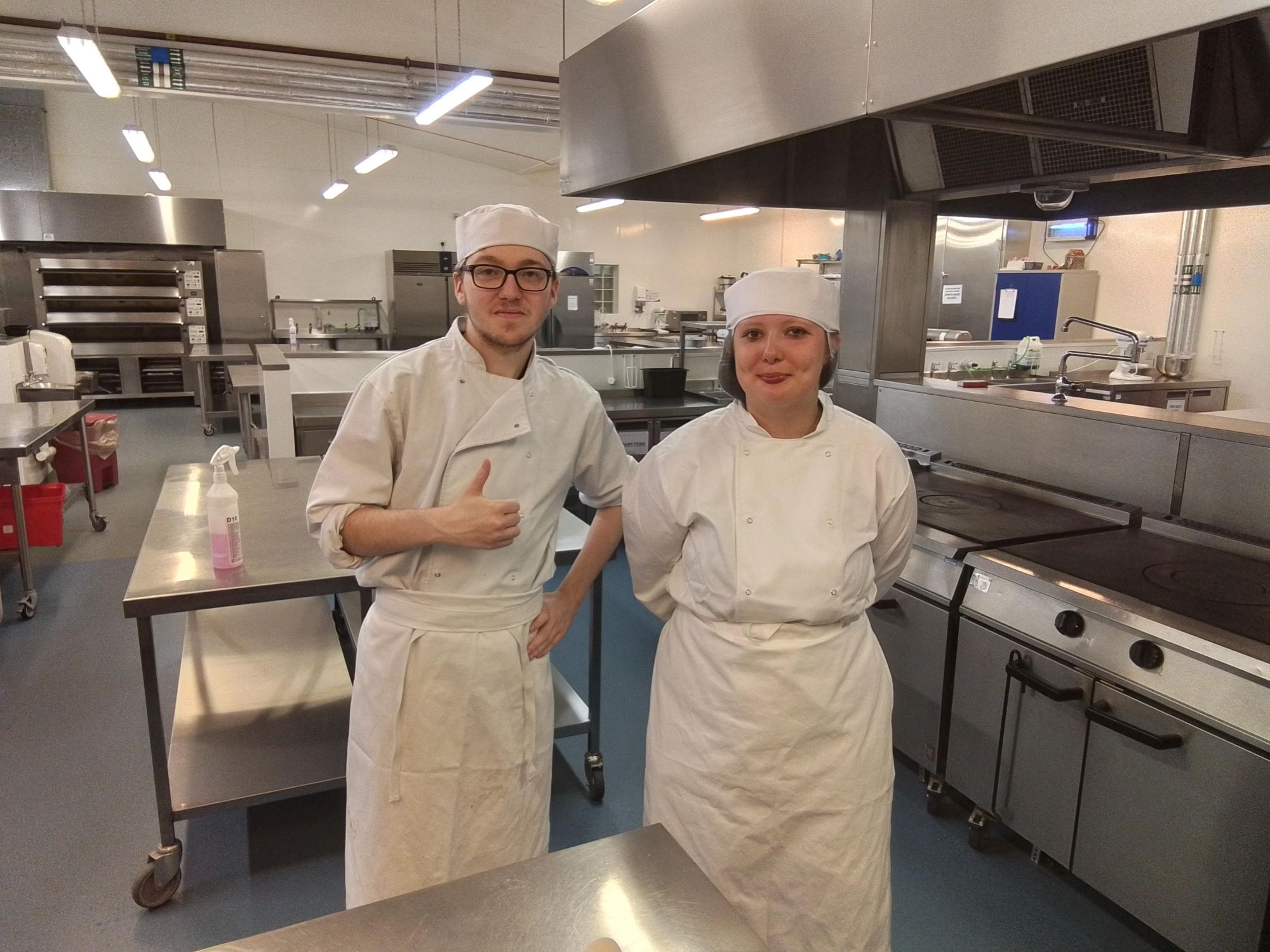 Two students in an undustrial kitchen. The students are dressed in chefs whites. The student on the left is wearing glasses and doing a thumbs up motion with their hand