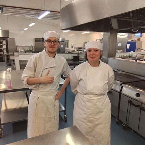 Two students in an undustrial kitchen. The students are dressed in chefs whites. The student on the left is wearing glasses and doing a thumbs up motion with their hand