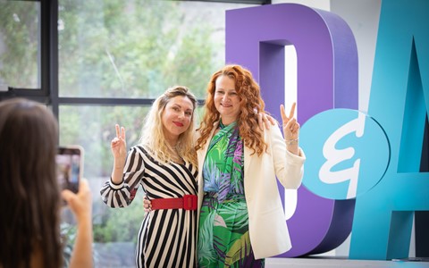 Two ESOL students standing in front of the giant D&A letters at Gardyne. One is blonde with a black and white striped dress and the other has red hair with a tropical print dress. They are both holding up peace signs with their hands