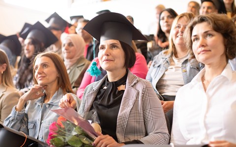 ESOL students sitting in an auditorium wearing graduation caps they made out of card. One holds a bouquet of flowers