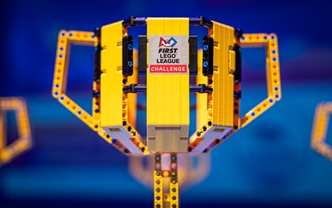 Close-up of a yellow LEGO trophy built from yellow and black LEGO pieces, featuring the 'FIRST LEGO League Challenge' logo. The trophy has a rectangular base and two side handles. The blurred background shows shades of blue and hints of other similar trophies