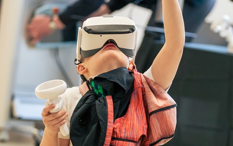 A child kneels on the floor wearing a virtual reality headset and holding two VR controllers with raised hands. A colourful jacket is draped over one arm. In the background, another person is working at a desk with various equipment.
