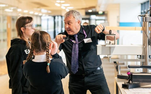 Three individuals gathered around a mechanical testing machine in a well-lit indoor setting. Two appear to be students in casual clothing, and the third, likely an instructor, wears a name tag and lanyard. The instructor is demonstrating or explaining the machine, which features clamps and levers. Papers are placed on a nearby table