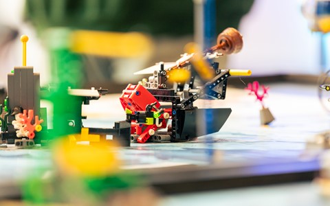 Close-up of a LEGO robotics competition setup featuring various LEGO structures and robots on a mat with green, red, black, and yellow elements. The background is blurred to emphasize the LEGO creations in the foreground