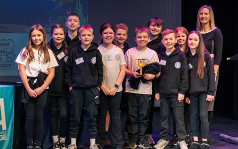 A group of children wearing matching black hoodies with logos and some with name tags stand on a stage next to an adult dressed in black. A table covered with a blue cloth is positioned to the left, and a banner or screen with partially visible D&A logo in the background. The student in the middle holds a yellow trophy made from lego.