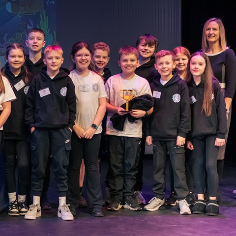 A group of children wearing matching black hoodies with logos and some with name tags stand on a stage next to an adult dressed in black. A table covered with a blue cloth is positioned to the left, and a banner or screen with partially visible D&A logo in the background. The student in the middle holds a yellow trophy made from lego.
