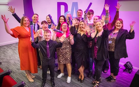 A group of people posing for a photo in a room with purple lighting. They are dressed in formal and semi-formal attire, with some holding rolled-up certificates or diplomas. Behind them is a large white wall with the letters 'DA' in purple and blue. Red chairs are visible on the left, and the floor is carpeted.