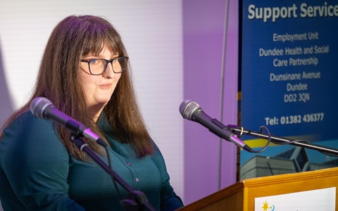 A person with long hair stands behind a podium, speaking into two microphones. Behind them is a banner displaying contact information for the Support Services Employment Unit, part of the Dundee Health and Social Care Partnership.