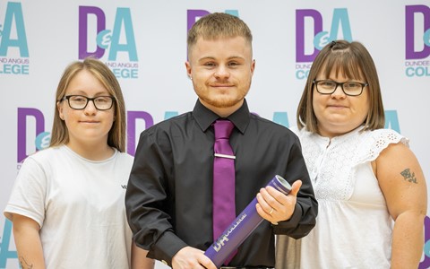 "Three people standing in front of a backdrop that repeatedly displays 'Dundee and Angus College.' The person in the center wears a black shirt with a purple tie and holds a purple certificate tube, while the two individuals on either side wear casual white tops