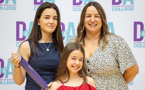 Three people standing in front of a backdrop that repeatedly displays 'Dundee and Angus College' with the letters 'D&A' in blue and purple. The girl on the left holds a purple certificate tube, the middle person is a child in a red outfit, and the woman on the right wears a patterned dress.