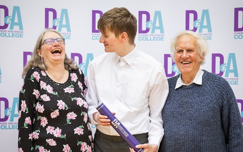 Three people standing in front of a backdrop that repeatedly displays 'Dundee and Angus College.' The person in the middle holds a rolled-up certificate or diploma with the same text.
