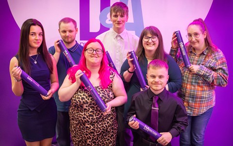 A group of seven students standing together, each holding a purple certificate tube. A large purple and white banner with the letters 'DA' is visible in the background. The individuals' faces are blurred, and they are dressed in a mix of formal and casual clothing.