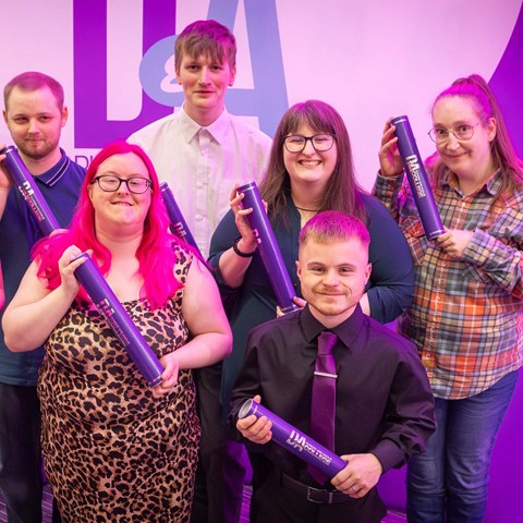 A group of seven students standing together, each holding a purple certificate tube. A large purple and white banner with the letters 'DA' is visible in the background. The individuals' faces are blurred, and they are dressed in a mix of formal and casual clothing.