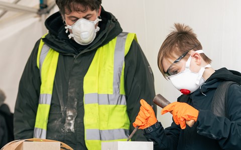 Two individuals working on a chiseling project. The person in the foreground, wearing orange gloves, is carving a design into a white block. Behind them, another person in a high-visibility yellow vest over a black jacket stands observing. The table holds another white block and various tools
