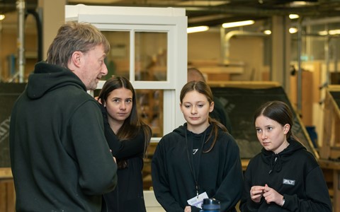 Four people gathered around a wooden workbench in a workshop. One person, standing on the left and wearing a black hoodie with tools on their belt, appears to be demonstrating or explaining something. The other three, also in black hoodies, are standing and listening. The workbench holds woodworking tools, a blue water bottle, and red ear protection. A window frame and workshop equipment are visible in the background