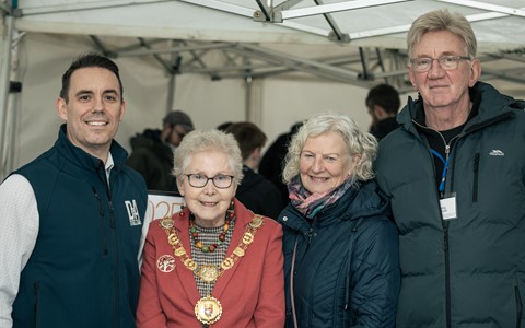 Four people standing together under a tent. The central figure wears a red outfit with a large gold chain and medallion, suggesting a ceremonial or official role. The other three individuals are dressed in casual outdoor clothing, including jackets and vests