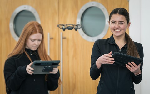 Two individuals standing indoors, each holding a tablet. The person on the left has long red hair and is looking down at their device, while the person on the right, with long dark hair tied back, is smiling. A small drone hovers in mid-air between them. Behind them are two wooden doors with round windows