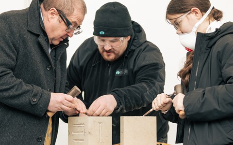Three individuals carving designs into two stone blocks. The person on the left, wearing glasses and a dark coat, uses a chisel and hammer on a block with two vertical lines. The middle person, in a black beanie and jacket, assists with the carving. The person on the right, with long hair tied back and wearing a face mask, also uses a chisel and hammer on an uncarved stone block