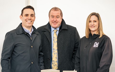 Three individuals standing behind a table displaying two concrete blocks. The person on the left wears a dark jacket over a light blue shirt. The middle person is dressed in a dark overcoat, blue shirt, and a patterned yellow tie. The person on the right wears a black jacket with 'DA' on it. The concrete blocks on the table have partially visible markings or letters etched into them