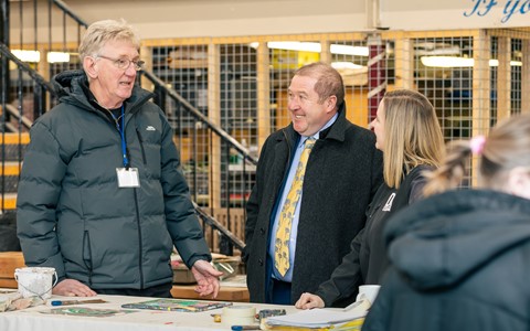 Four people gathered around a table with art supplies in an indoor setting. One person wears a black jacket with a name badge, another is in a suit with a yellow tie, and a third wears a black hoodie. A fourth person is partially visible in the foreground. The background includes stairs and metal railings
