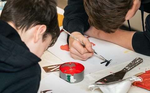 Two individuals seated at a table engaged in painting. One is using a thin brush and black paint to create a design on paper, while the other works with red and black paint on a separate sheet. The table is scattered with art supplies, including a color palette, masking tape, a green pencil, and containers of red and green paint.