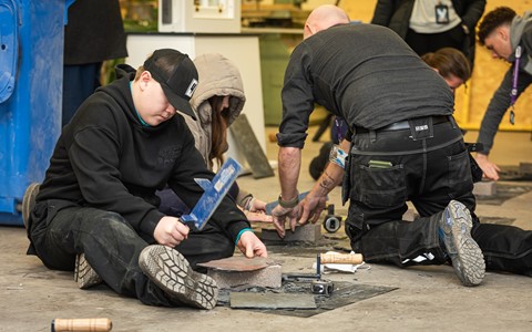 Four individuals engaged in a stone or tile cutting project. In the foreground, one person sits on the ground wearing a black hoodie and cap, using a hammer and chisel. Another person kneels nearby, also working with tools on a stone or tile. Various tools, including hammers and chisels, are scattered around. In the background, two more people are involved in similar activities.