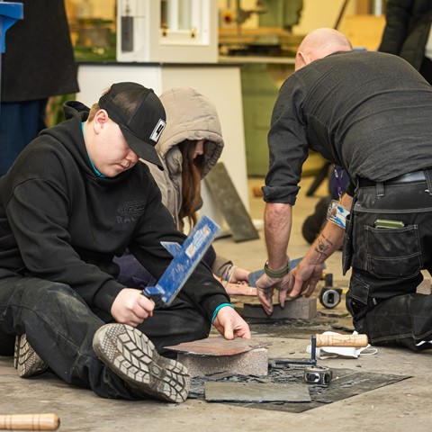 Four individuals engaged in a stone or tile cutting project. In the foreground, one person sits on the ground wearing a black hoodie and cap, using a hammer and chisel. Another person kneels nearby, also working with tools on a stone or tile. Various tools, including hammers and chisels, are scattered around. In the background, two more people are involved in similar activities.