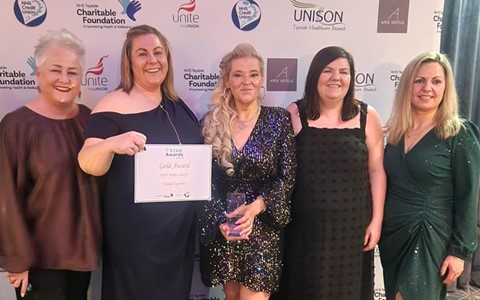 Five women standing together at an awards event, dressed in formal attire. They are posing in front of a backdrop featuring sponsor logos such as 'Unite,' 'UNISON,' and 'Apex Hotels.' One woman holds a certificate labeled 'Gold Award,' and another holds a trophy