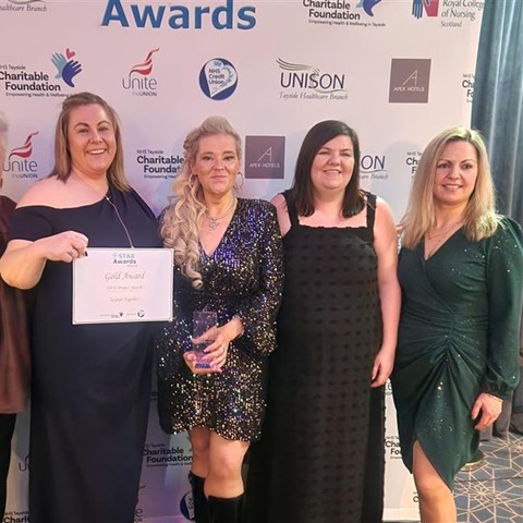 Five women standing together at an awards event, dressed in formal attire. They are posing in front of a backdrop featuring sponsor logos such as 'Unite,' 'UNISON,' and 'Apex Hotels.' One woman holds a certificate labeled 'Gold Award,' and another holds a trophy