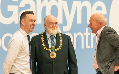 Three men standing and conversing in front of a blue background with partially visible white text reading 'Gardyne Sports Centre.' The man in the middle wears a suit, tie, and a ceremonial chain, while the other two are dressed in business attire.