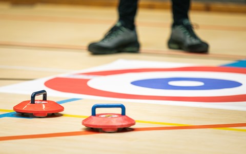 Close-up of two red curling stones with blue handles on an indoor curling rink. The stones are near the target area, marked with blue, white, and red concentric circles. A person's legs and feet in black shoes are visible in the background
