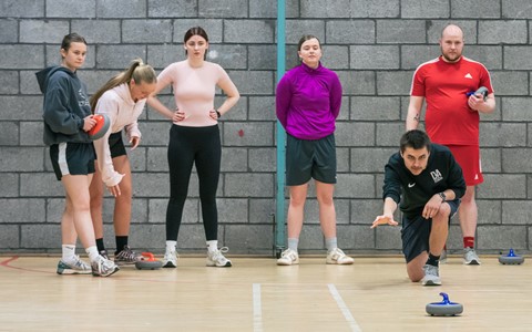 Six people in a gymnasium with a brick wall background. One person, dressed in black, is kneeling on the floor and is playing a game of curling. The other five individuals are standing and observing, some holding curling stones.