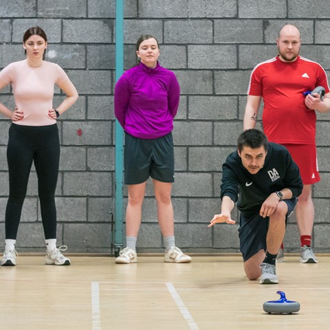 Six people in a gymnasium with a brick wall background. One person, dressed in black, is kneeling on the floor and is playing a game of curling. The other five individuals are standing and observing, some holding curling stones.