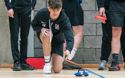 A group of people in a gymnasium. One person, kneeling on the floor in a black hoodie and shorts, throws a blue curling stone. Other individuals stand nearby, some holding red curling stones. A grey brick wall is visible in the background.