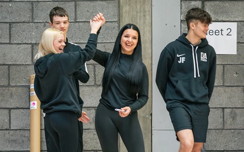 Four people standing in a gymnasium. On the left, two individuals are holding hands—one in black athletic wear and the other in a black hoodie and leggings. On the right, two others stand side by side, both wearing black hoodies, with one also in shorts. A sign on the wall reads 'Sheet 2