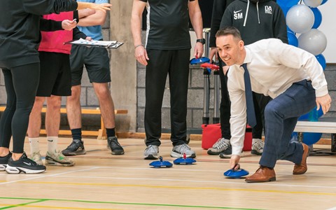 "A group of people in a gymnasium. One person, wearing a white shirt, blue tie, and brown shoes, is crouching and placing a blue curling stone on the floor. Others stand nearby, observing or holding equipment. A stack of balloons is visible in the background on the right."