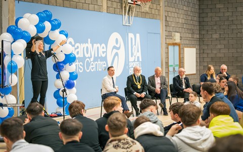 A speaker stands on the left, gesturing with one hand and holding a microphone, under an arch of blue and white balloons. Behind them, a blue wall displays the text 'Gardyne Sports Centre'. On the right, five individuals are seated facing an audience, which consists of people sitting on the floor. The setting is an indoor sports hall with a basketball hoop in the background.