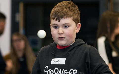 A child wearing a black hoodie with the text 'STRONGER THAN I THINK,' is playing table tennis. The boy is holding a paddle and appears to be engaged in the game. The background is slightly blurred, showing other people present in the indoor setting, but they are not in focus