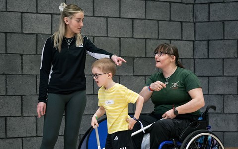"A young boy in a yellow striped shirt and black shorts stands on an indoor court holding a sports stick. He is accompanied by two adults—one standing and one in a wheelchair, who is also holding a sports stick. A grey brick wall is visible in the background.