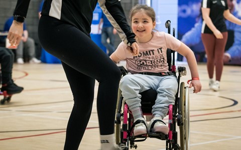 A young girl in a wheelchair playing with a soccer ball in an indoor gymnasium. An adult in sports attire stands beside her, appearing to assist. In the background, other people are engaged in various activities, and a banner is visible on the wall