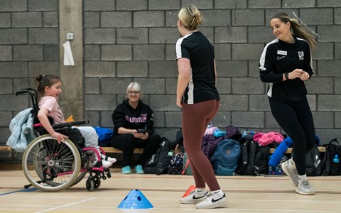 A gymnasium scene with four people. In the foreground, two women—one in a black shirt and maroon leggings, the other in a black tracksuit—are engaged in an activity involving blue and orange cones on the floor as a young girl in a wheelchair watches. Another person sits on the floor against a brick wall surrounded by bags.