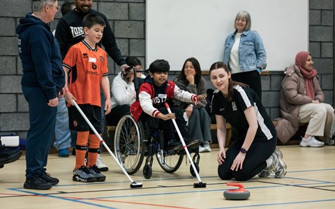 A group of people in an indoor sports facility. In the foreground, a child in a wheelchair wearing a red jacket and glasses holds a curling broom, assisted by another child in an orange sports uniform and an adult in a black shirt with white sleeve stripes. A curling stone is on the floor nearby. In the background, several people are seated or standing against a wall, observing the activity