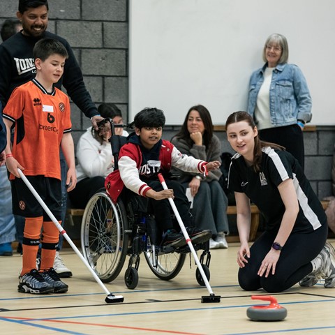 A group of people in an indoor sports facility. In the foreground, a child in a wheelchair wearing a red jacket and glasses holds a curling broom, assisted by another child in an orange sports uniform and an adult in a black shirt with white sleeve stripes. A curling stone is on the floor nearby. In the background, several people are seated or standing against a wall, observing the activity