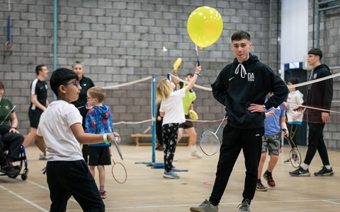 An indoor gymnasium with a brick wall background, where a mixed group of children and adults are actively participating in a badminton session. Several badminton nets are set up across the gym, and the floor is marked with white court lines. Some participants are holding badminton rackets mid-play, while others are standing or walking around. One individual is holding a large yellow balloon, possibly as part of an adapted activity. The scene is lively and inclusive, with people spaced out across