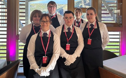 Six individuals dressed in formal attire—white shirts, black aprons, and red lanyards with badges—stand inside a room with large windows. The windows offer a view of mountains and greenery outside. The individuals' faces are blurred. Tables in the room are covered with white tablecloths, and one table displays a sign with the number '2'