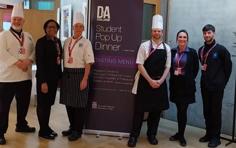 Six people standing in a line on a wooden floor, posing next to a tall banner that reads 'D&A College Student Pop Up Dinner Tasting Menu.' Some individuals are dressed in chef uniforms, while others wear black attire. The setting features a modern interior with light-coloured walls and large windows