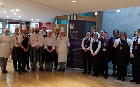 A group of chefs and waitstaff stand together in a well-lit room with wooden floors. The chefs are wearing white uniforms and hats, while the waitstaff are dressed in black vests and white shirts. In the center of the group is a tall banner that reads 'DA Student Pop-Up Dinner' with additional smaller text below it. The background includes large windows and modern decor elements